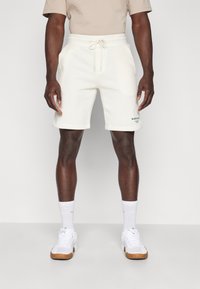 Weiße Baumwollshorts mit einem elastischen Bund und Kordelzug, versehen mit einem kleinen schwarzen Logo-Detail am linken Bein. Modell kombiniert mit weißen Schuhen und Socken.