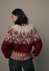 Chunky gebreide trui met een rood en roomkleurig kleurenschema, voorzien van sneeuwvlokpatronen, geribbelde randen en een hoge, gestructureerde halslijn.