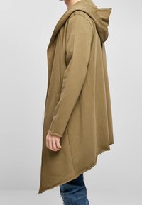 Larga cárdigan con capucha en color verde oliva y ajuste relajado. Presenta una tela texturizada, frente abierto y dobladillo deshilachado. Lleva puesta sobre unos jeans casuales.