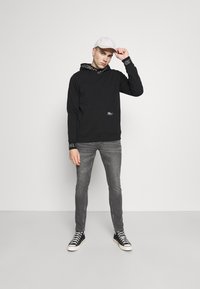 Hoodie preto com um pequeno logotipo, jeans cinzentos e sapatilhas pretas com solas brancas. O modelo usa um boné de cor clara e exibe uma pose descontraída.