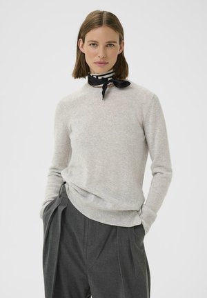 Maglione grigio chiaro con scollo rotondo e polsini a coste, abbinato a pantaloni grigio scuro a pieghe e una sciarpa a motivo bianco e nero.