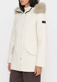 Giacca bianca imbottita con cappuccio foderato in pelliccia, chiusura frontale con cerniera, ampie tasche laterali e una toppa nera con logo sulla manica.