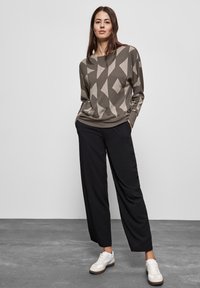 Sweatshirt kaki avec un motif géométrique, associé à un pantalon large noir et des sneakers blanches, sur fond neutre.