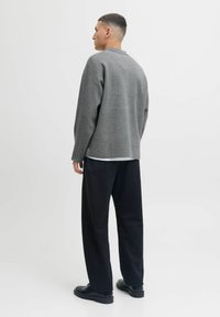 Muškarac stoji okrenut lijevo, odjeven u sivi duksericu dugih rukava, crne hlače širokog kroja i crne cipele, na jednostavnoj bijeloj pozadini.