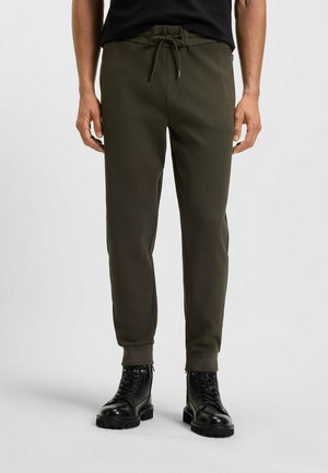 Dunkelgrüne Jogginghosen mit einem elastischen Bund, Kordelzugverschluss und gerippten Bündchen. Kombiniert mit schwarzen Schnürstiefeln.