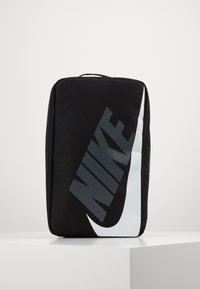 Svart rektangulär Nike-väska med stor vit swoosh-logotyp och mörk "NIKE"-text, stående upprätt på en vit yta mot en enfärgad bakgrund.