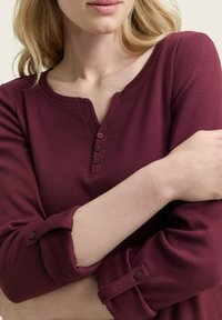 Frau in einem langärmligen bordeauxroten Ripp-Top mit drei Knöpfen am Ausschnitt und hochgerollten Ärmeln, die die Arme vor der Brust verschränkt.