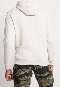 Krémová mikina s kapucí na šňůrku, žebrovanými manžetami a spodním lemem, doplněná o maskáčové cargo kalhoty. Hladká textura a uvolněný střih.