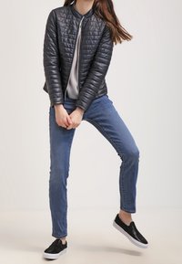 Kobieta w czarnej pikowanej kurtce, w pasiastej koszulce, niebieskich dżinsach i czarnych butach na slip-on, stoi z nogami rozstawionymi i z dłońmi złożonymi.