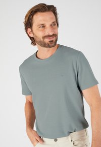 Camiseta de manga corta y cuello redondo en un tono verde claro, con una tela texturizada y un sutil detalle del logo en el pecho.