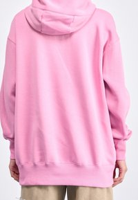 En person som bär en lös rosa hoodie och beige byxor, sedd bakifrån mot en vit bakgrund.