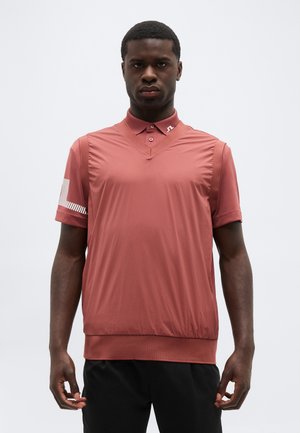 Noore mehega tume nahaga, kes kannab tolmroosat lühikeste varrukatega polo särke kraega ja musti pükse, seisab heleda tausta ees.