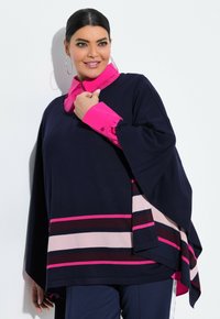 Frau trägt einen marineblauen Poncho mit pink- und beige-gestreiften Mustern über einem leuchtend pinken Hemd, dunklen Hosen und großen Creolen.