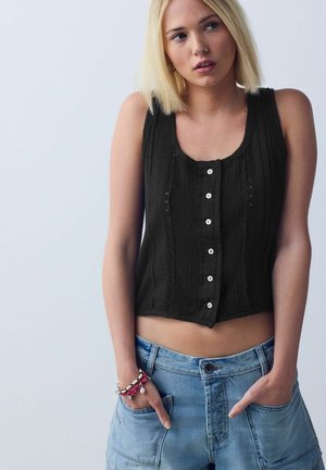 Young woman with short blonde hair wearing a black sleeveless buttoned top and blue jeans, hands in pockets, against a plain background.