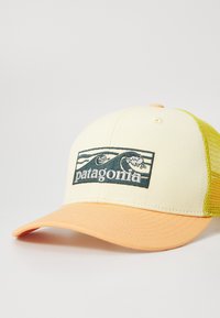 Patagonia-Trucker-Cap mit einer cremefarbenen Vorderseite, gelbem Netzrücken und orangenem Schirm. Die Vorderseite zeigt ein gesticktes Logo mit Wellen und Text.