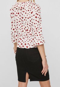 Blusa bianca con motivo a cuori rossi, colletto volant e maniche a sbuffo, abbinata a una gonna di jeans nera con spacco posteriore.