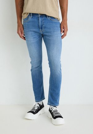 Personne portant un jean slim bleu clair, une chemise beige et des baskets épaisses noires et blanches, debout sur un fond blanc uni.