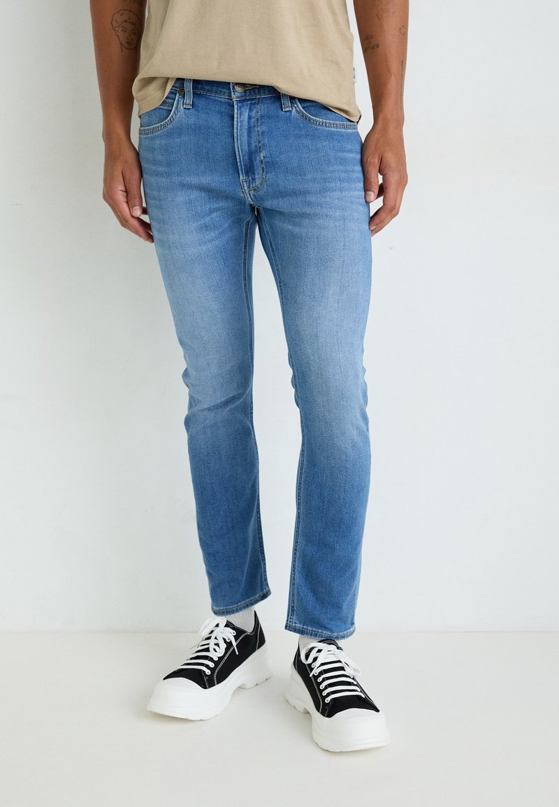 Personne portant un jean slim bleu clair, une chemise beige et des baskets épaisses noires et blanches, debout sur un fond blanc uni.