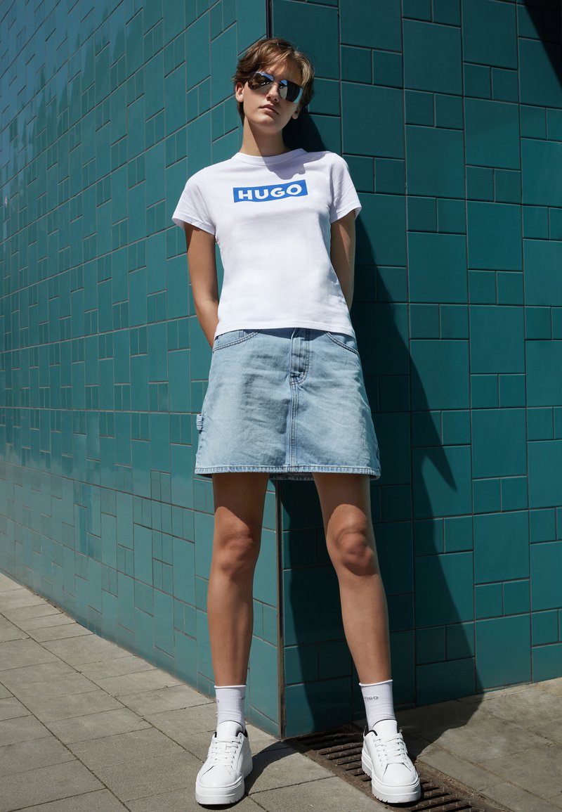 Bílý bavlněný tričko s modrým logem "HUGO", zkombinováno s světlou džínsovou sukní. Modelka nosí bílé tenisky a sluneční brýle, stojí před tyrkysovou zdí.