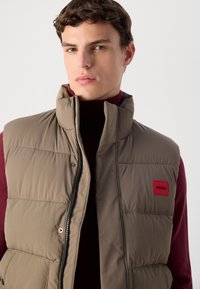 Young man with short brown hair wearing a taupe padded vest over a burgundy turtleneck against a plain background.