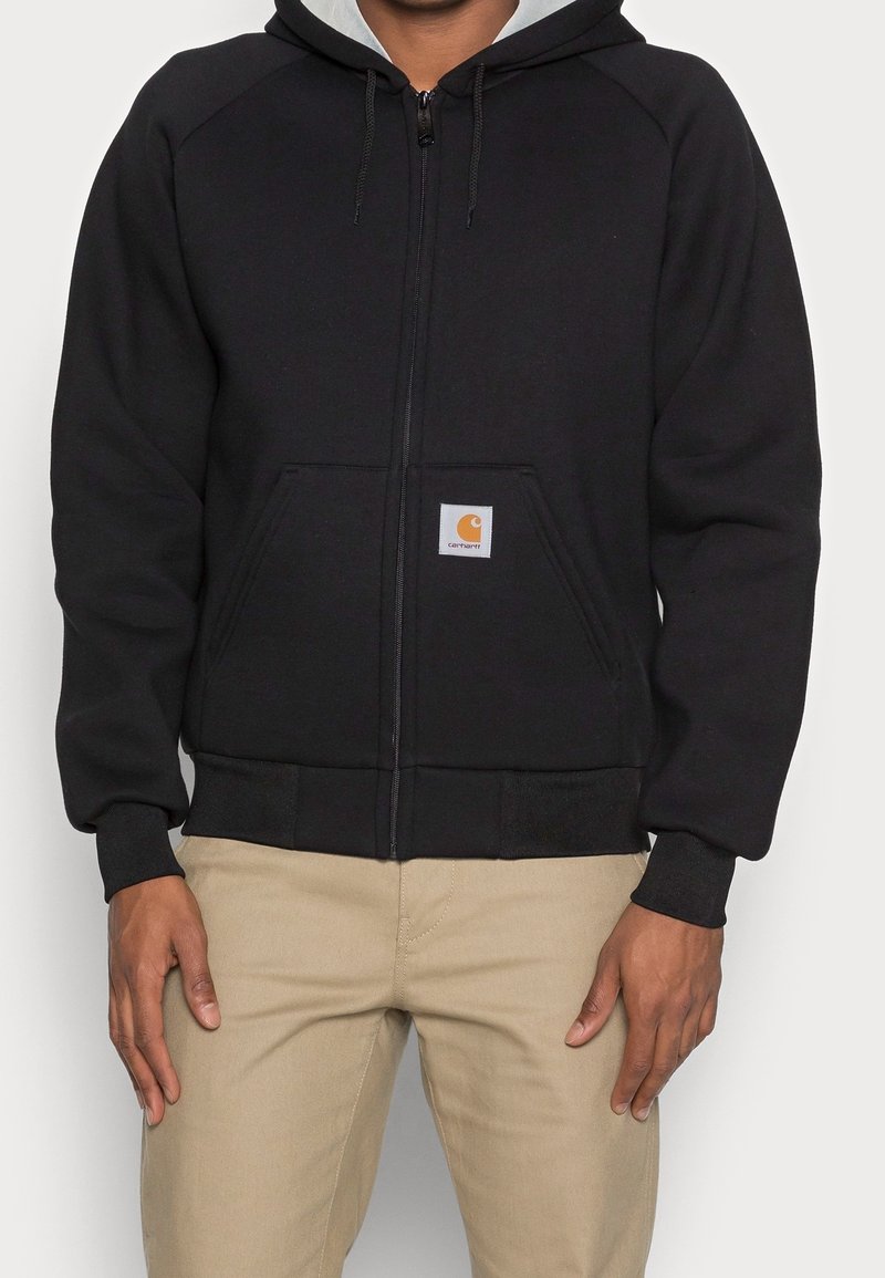 Uomo che indossa una felpa nera con zip e tasche frontali, insieme a pantaloni beige, mostrato dalla parte centrale del torso ai fianchi su uno sfondo neutro.