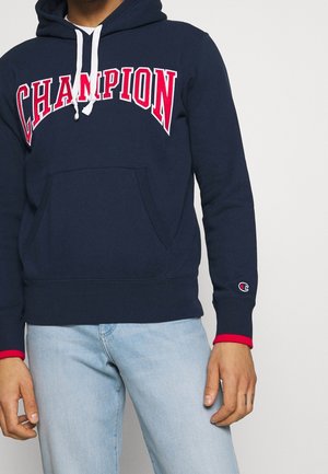 Persona che indossa una felpa con cappuccio Champion blu navy con logo rosso e bianco e jeans azzurri, in piedi davanti a uno sfondo semplice.
