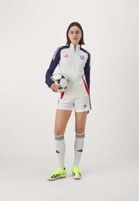 Top deportivo blanco y azul marino con acentos rojos, shorts blancos y calcetas grises. Botas de fútbol amarillas. Sosteniendo un balón de fútbol en blanco y negro.