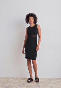 Vestido preto sem mangas com uma silhueta ajustada, apresentando um painel de cintura transparente. Modelo combinado com sandálias pretas grossas. Textura suave.