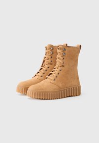 Bottines en daim tan à lacets avec semelles épaisses côtelées et languettes de tirage, conçues pour un usage décontracté sur un fond gris neutre.