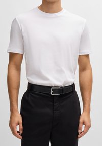 Biała bawełniana koszulka w połączeniu z czarnym skórzanym paskiem z prostokątną srebrną klamrą, noszona z ciemnymi spodniami.