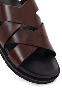 Close-up van een zwarte sandalenzool met brede, donkerbruine leren bandjes die over het voetgedeelte kruisen, zichtbare gestikte rand van de zool.