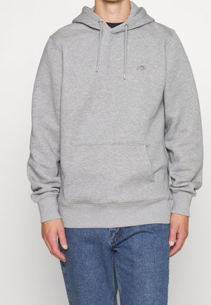 Sudadera gris con un bolsillo frontal, capucha con cordón, puños acanalados y dobladillo. Presenta un pequeño logo en el pecho. Lleva puestas unas jeans de mezclilla.