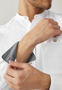 Bílá lněná košile s stojatým límečkem a šedými ohrnutými manžetami. Manžeta má jemnou texturu a ozdobný knoflík.