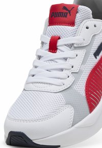 Białe sportowe buty z siateczkową cholewką, czerwonymi akcentami i szarymi sekcjami. Posiadają teksturowaną podeszwę i wyraźny system sznurowania. Widoczna jest również logo PUMA.