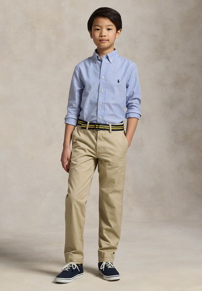 Chłopiec w niebieskiej paskowanej koszuli na guziki, beżowych spodniach z paskiem w paski i granatowych trampkach stoi z jedną ręką w kieszeni.