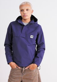 Veste à capuche de couleur violet profond, fabriquée en matériau résistant à l'eau. Elle dispose d'un col haut, d'une poche à l'avant et de poignets élastiques pour un ajustement près du corps.