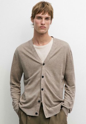 Junger Mann mit hellbraunen Haaren, der eine beigefarbene, geknöpfte Strickjacke über einem weißen Hemd und braune Hosen trägt und vor einem einfarbigen Hintergrund steht.