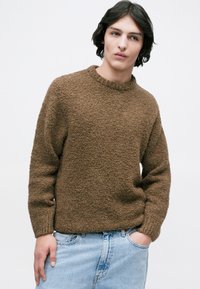 Rjav pulover z teksturo, okroglim izrezom in dolgimi rokavi, v kombinaciji s svetlo modrimi jeans hlačami. Tkanina ima mehak, puhast videz.