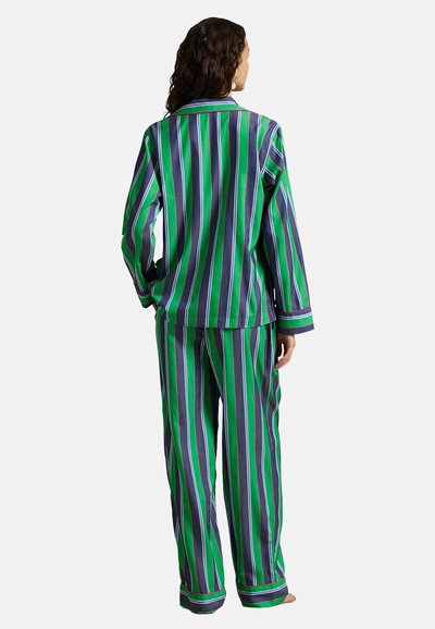 Frau steht barfuß, trägt einen grün-blau-weißen, vertikal gestreiften Pyjama mit langärmligem Oberteil und Hose, zeigt den Rücken vor weißem Hintergrund.