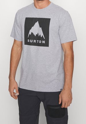 Mężczyzna noszący jasnoszary t-shirt Burton z czarnym logo góry oraz czarne spodnie z kieszenią naszytą na prawej udzie.