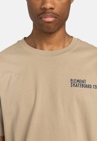 Beige Baumwoll-T-Shirt mit kurzen Ärmeln, das auf der linken Seite ein navy-blaues "ELEMENT SKATEBOARD CO."-Grafikdesign trägt. Glatte Textur.