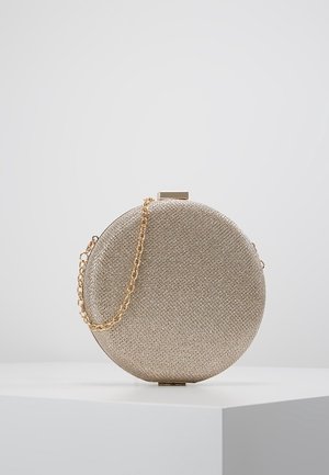 Rund gylden tekstureret clutch taske med kort gylden kædehåndtag på en hvid pedestal mod en lysegrå baggrund.
