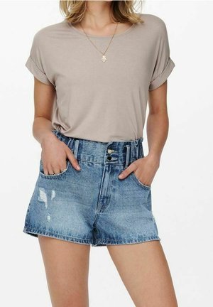Mujer con camiseta beige de mangas remangadas metida dentro de shorts de mezclilla azules de talle alto y desgastados, con las manos en los bolsillos.