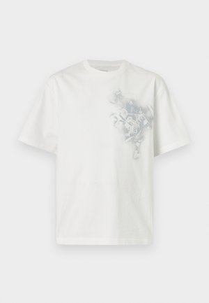 Homme portant une casquette noire, un T-shirt blanc avec un motif floral clair, un pantalon large à motif feuilles vertes et des chaussures noir et blanc, debout droit.