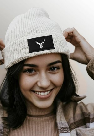 White knit beanie with a black patch featuring a deer head. The beanie has a ribbed texture and is worn slightly slouched.