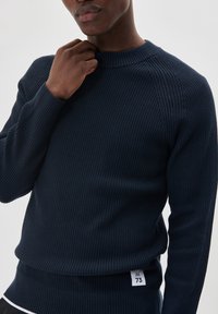 Navy ribbed knit sweater with a rounded neckline and long sleeves. Features a tag with "M 73" on the lower seam. Fabric appears textured.