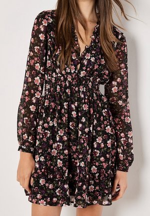 Femme portant une robe noire fleurie à manches longues, avec un col à volants et une taille froncée, debout devant un fond clair uni.
