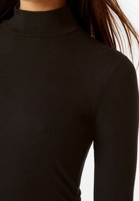 Haut col roulé noir côtelé avec une coupe ajustée. Le tissu est texturé et le col est haut, s'étendant jusqu'aux épaules.