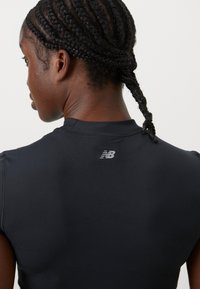 Haut de sport noir à manches courtes, texture lisse et logo réfléchissant New Balance dans le dos. Gros plan sur l'épaule et l'encolure.
