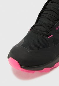 Czarne buty sportowe z różowymi akcentami, z siateczkową górą, usztywnionym noskiem i teksturowaną podeszwą dla lepszej przyczepności. Sznurowadła są starannie zawiązane.